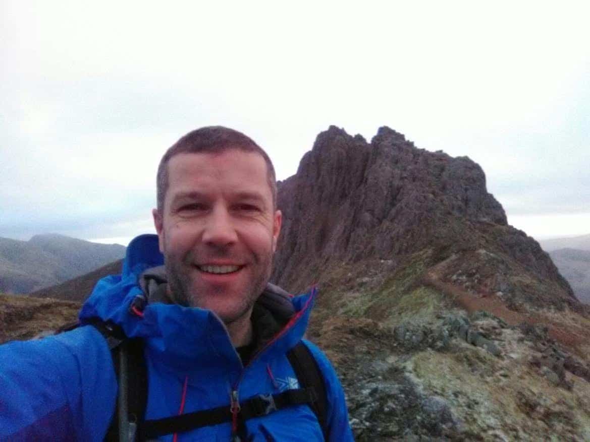 Man smiling in front of a rocky mountain peak.
