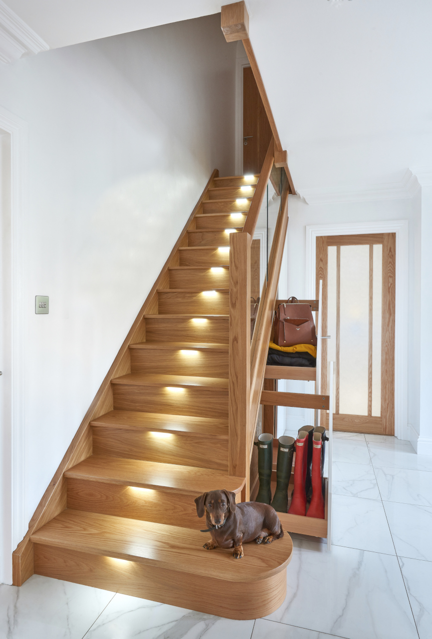 A small dog sits on the bottom step of a wooden staircase with built-in recessed lighting and storage unit, designed for boots and bags.
