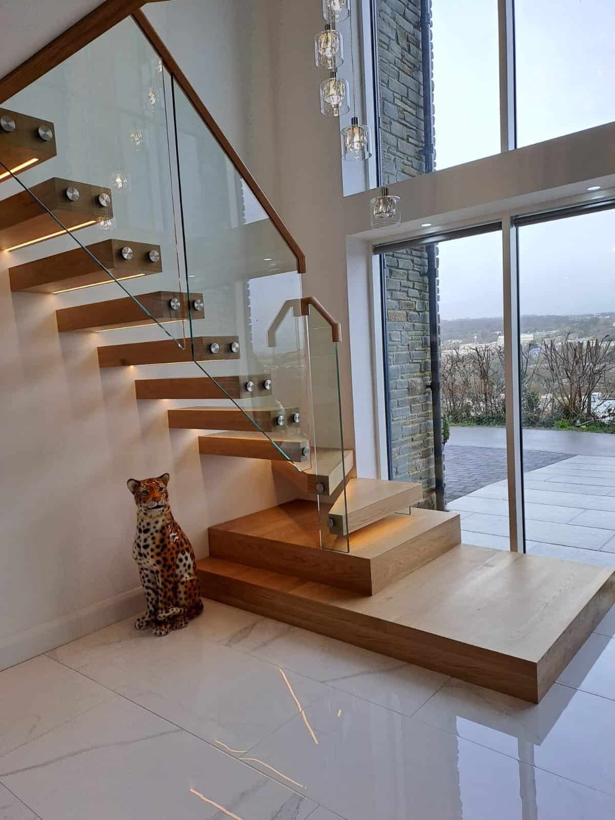 Floating oak staircase with a glass balustrade, wooden handrail and broad feature steps beside full-height windows.