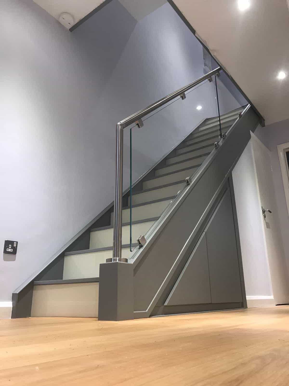 Grey staircase with glass balustrades, polished metal handrails and fitted cupboards beneath the stairs.