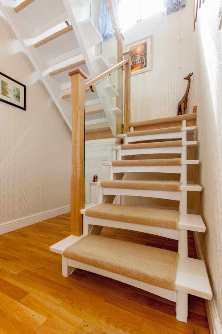 White open-riser staircase with beige carpet runners, oak newel posts and bracketed glass balustrades in a bright wood-floored hallway.