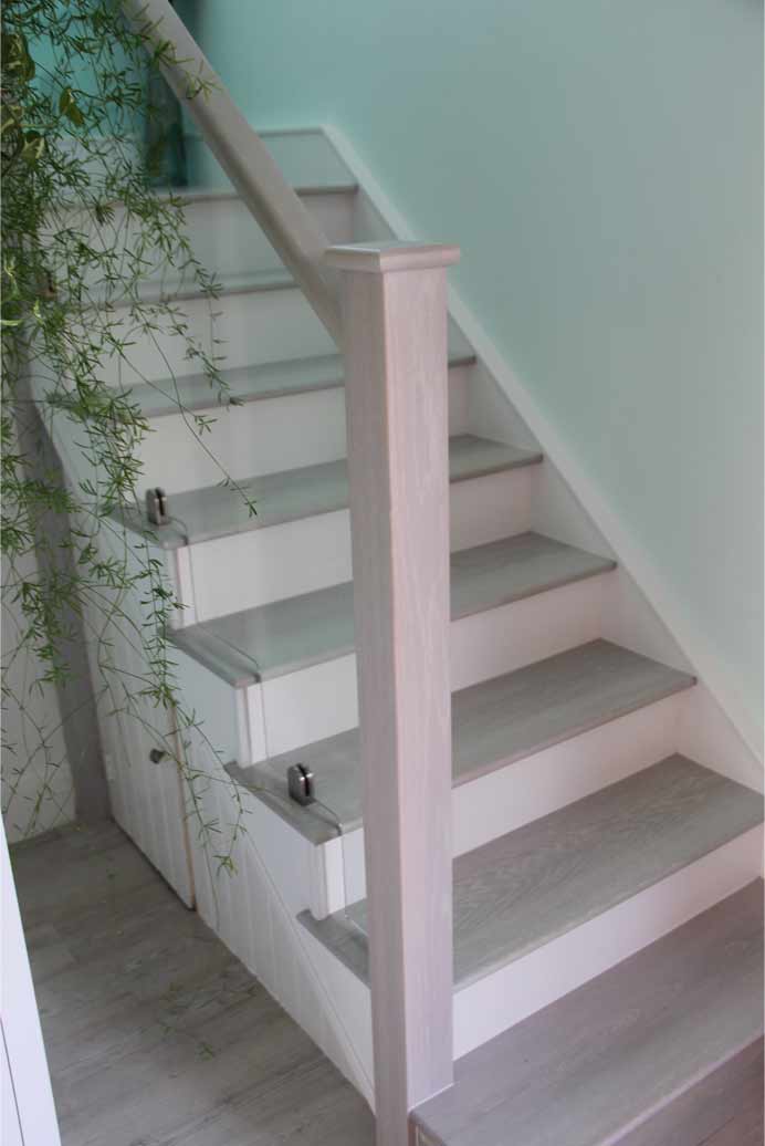 Pale painted staircase with grey wooden treads, a square newel post and glass panels secured by metal brackets beside a trailing plant.