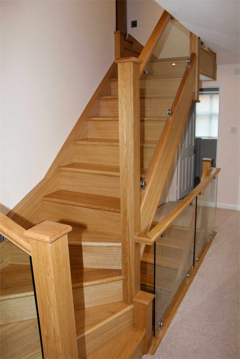 Turning oak staircase with wedge-shaped wooden treads, tall square posts and bracketed glass panels continuing around the stairwell.