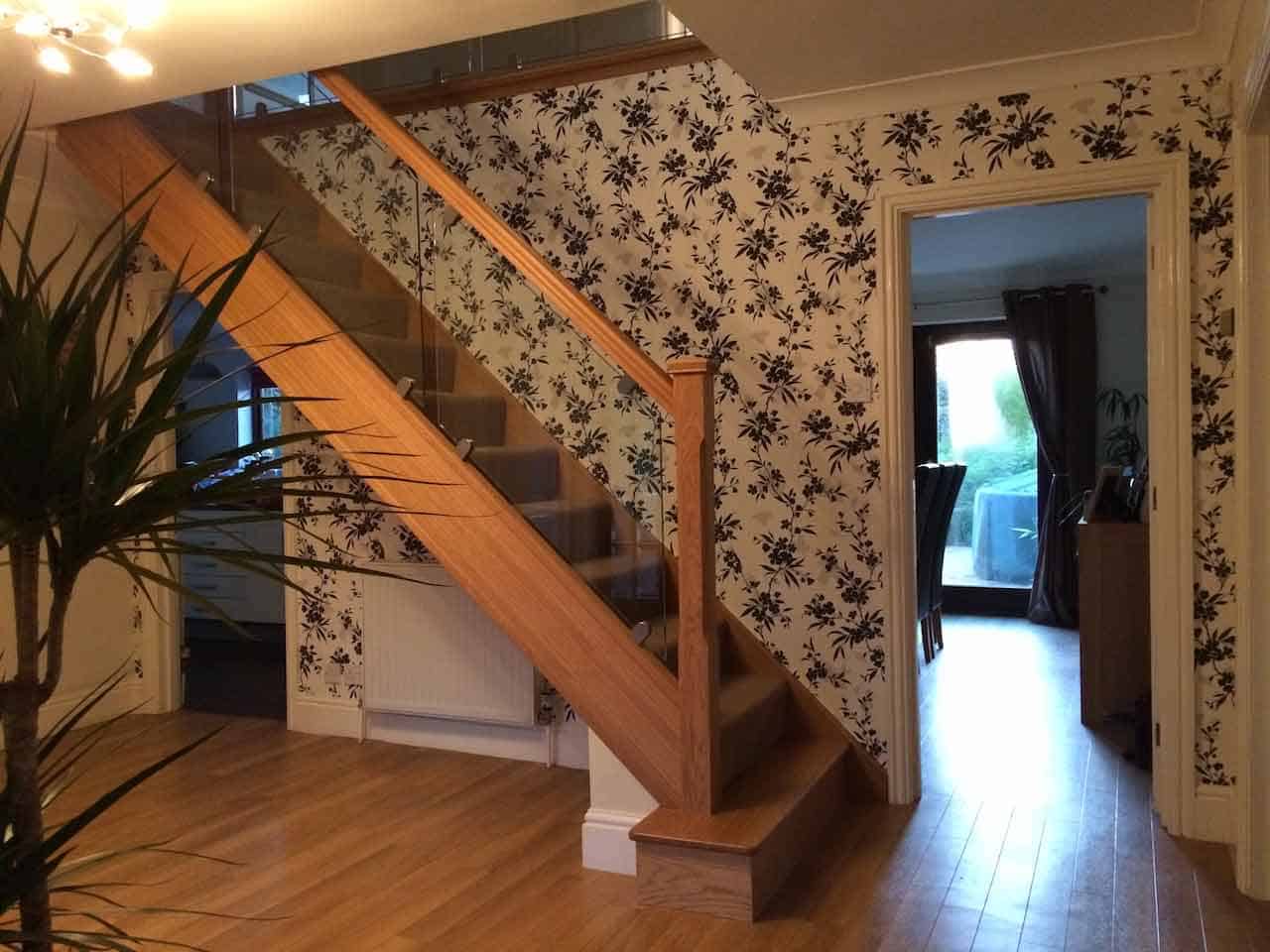 Carpeted oak staircase with clear glass balustrades and a wooden handrail beside black floral wallpaper in a wood-floored hallway.