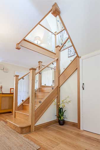 Oak staircase with bracketed glass panels, square newel posts and a rounded wooden bottom step beneath a matching glass gallery landing.