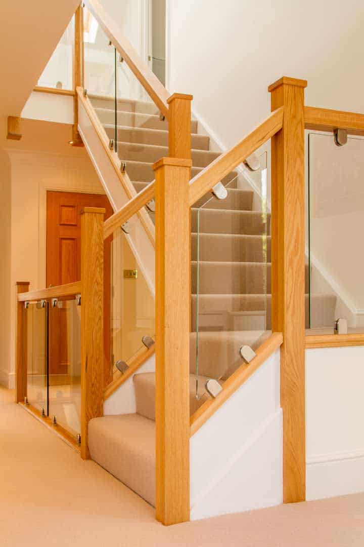 Cream carpeted staircase with white stringers, square oak newel posts and clear glass balustrades held by metal brackets across several levels.