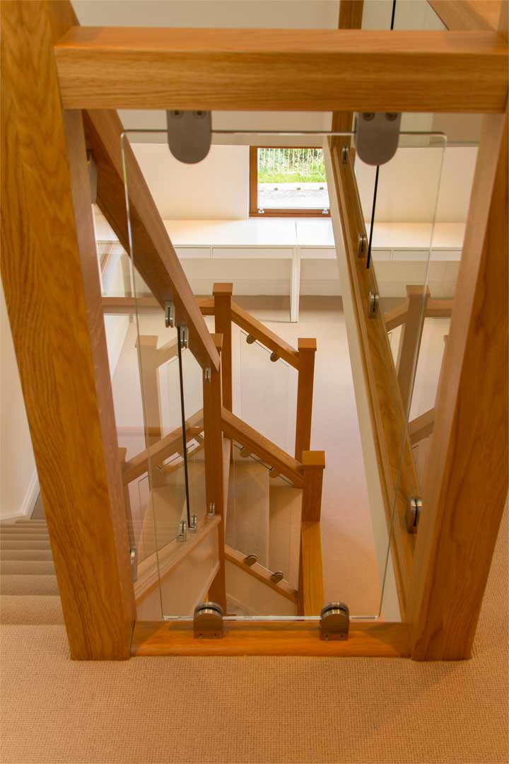 Close-up of clear landing glass secured by metal brackets within an oak frame, with matching glass stair balustrades visible below.