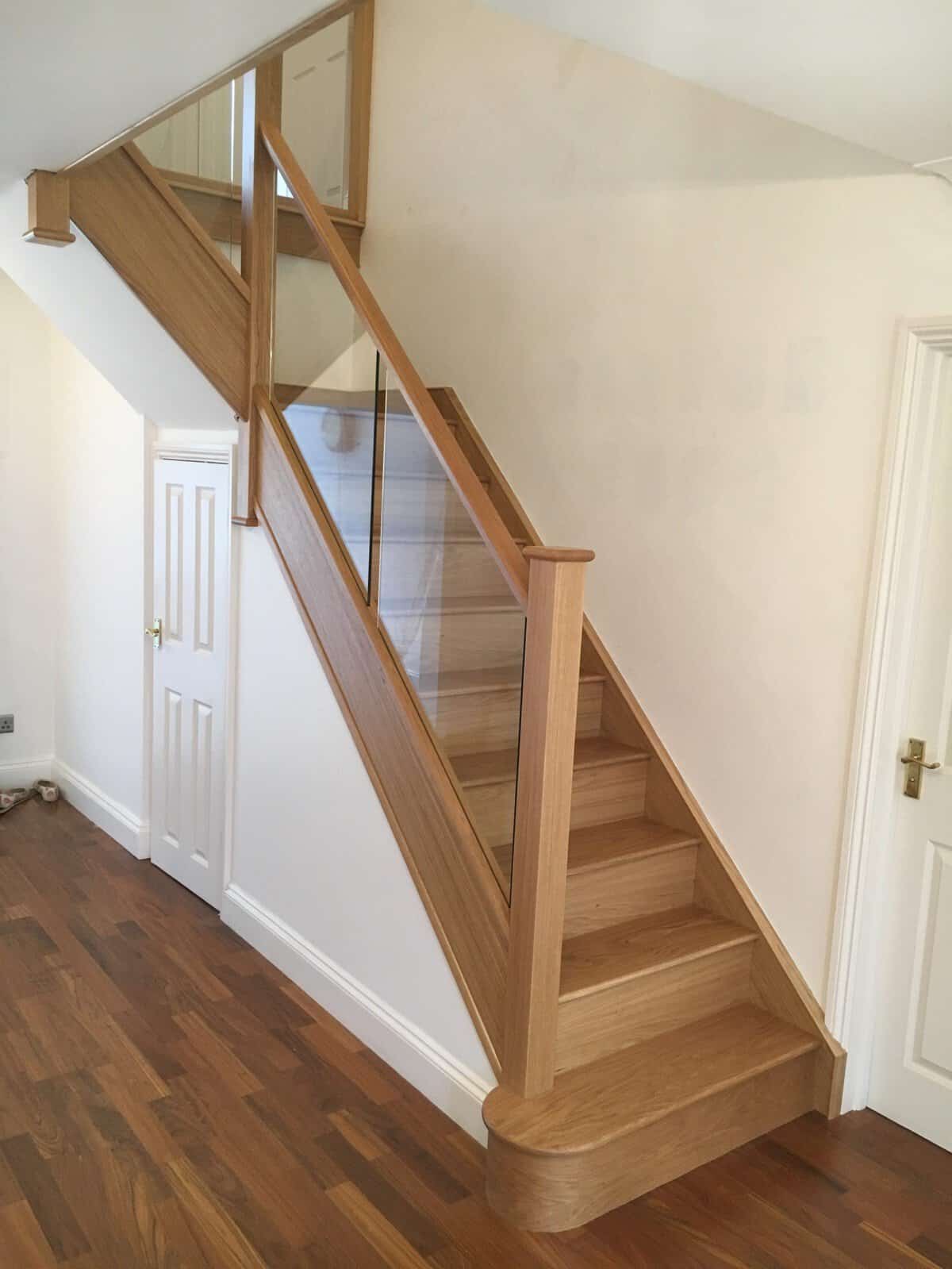 Straight oak staircase with clear glass balustrades, square wooden posts and a rounded bottom step above dark parquet flooring.