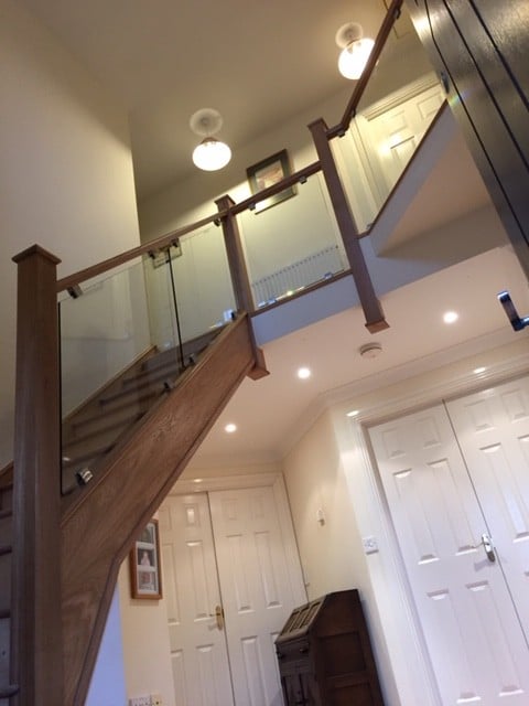 Low-angle view of an oak staircase and gallery landing with clear glass panels secured by metal brackets between square wooden posts.