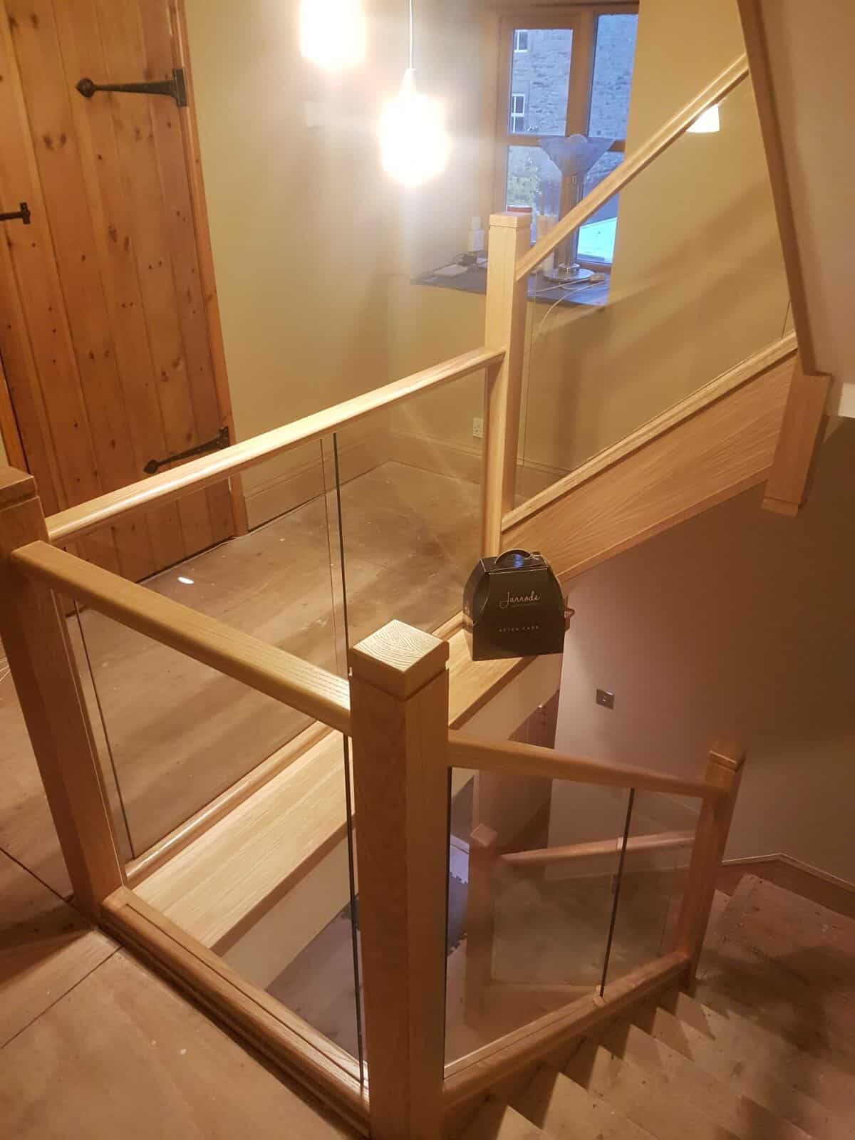 View down a turning oak staircase with clear glass balustrades, square wooden posts and matching rails around a tiled intermediate landing.