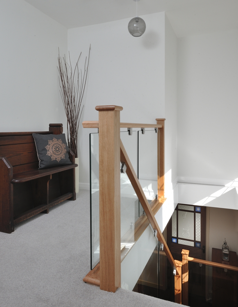 Sunlit carpeted landing with natural oak newel posts, matching handrails and clear glass panels secured by metal brackets around the stairwell.