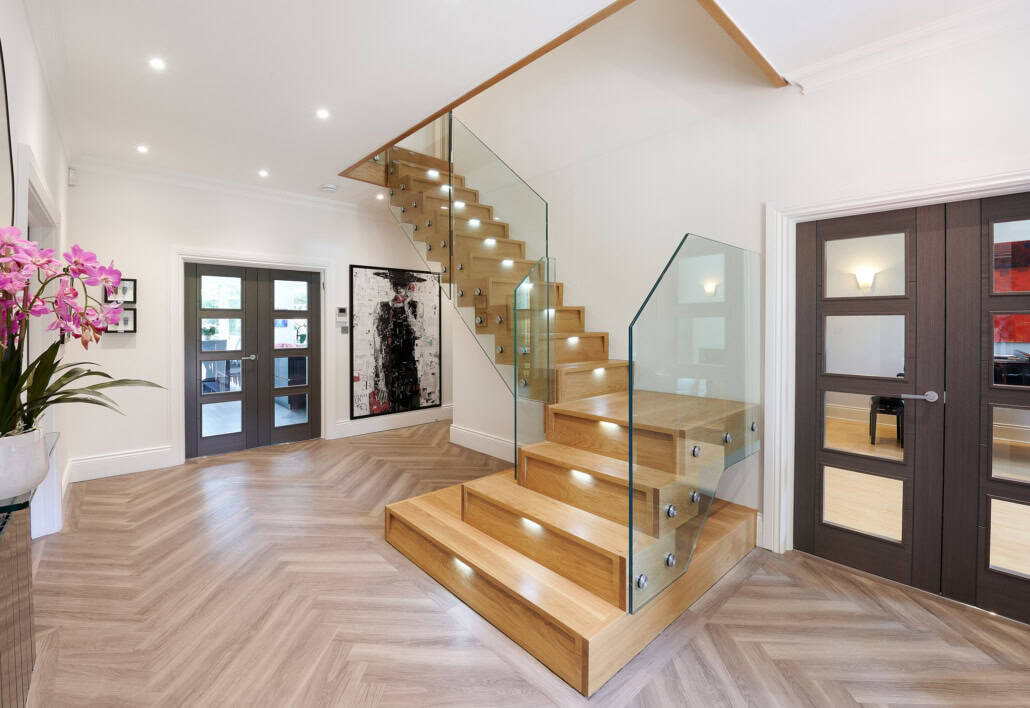 Oak staircase with recessed step lights and frameless glass balustrades secured with round metal fixings in a herringbone-floored hallway.