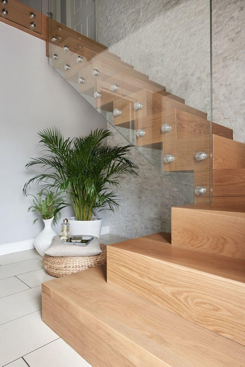 Oak staircase with closed risers and frameless glass balustrades secured by round metal fixings beside a pale stone feature wall.