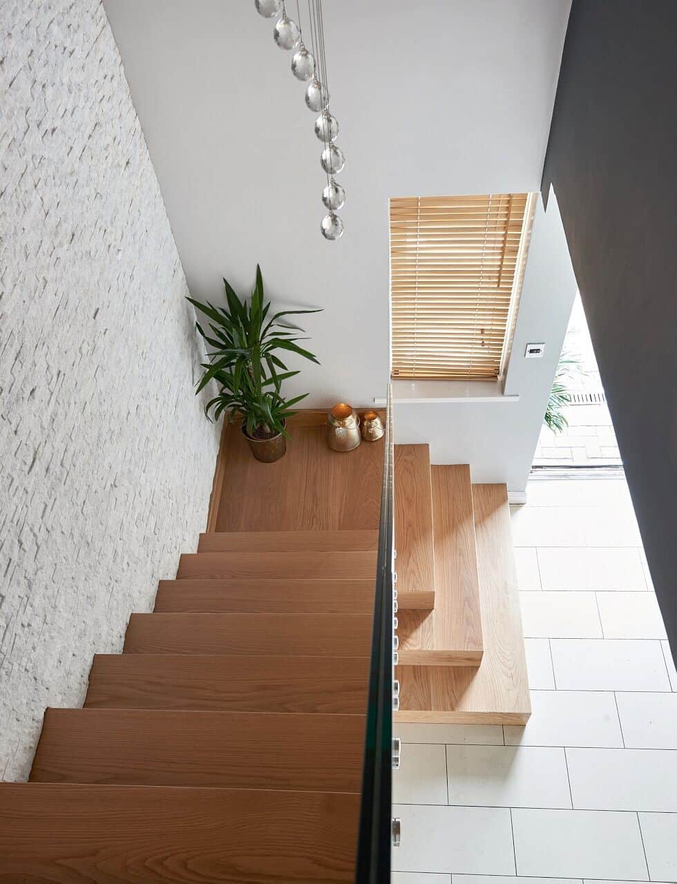 View down an oak staircase with a clear glass balustrade, turning lower steps and a stone feature wall beneath pendant lights.