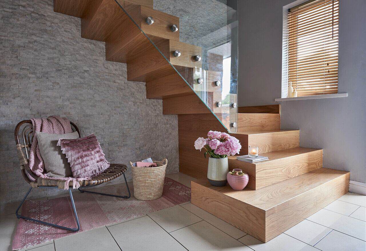 Turning oak staircase with a zigzag underside, broad lower steps and a frameless glass balustrade beside a stone feature wall and reading chair.