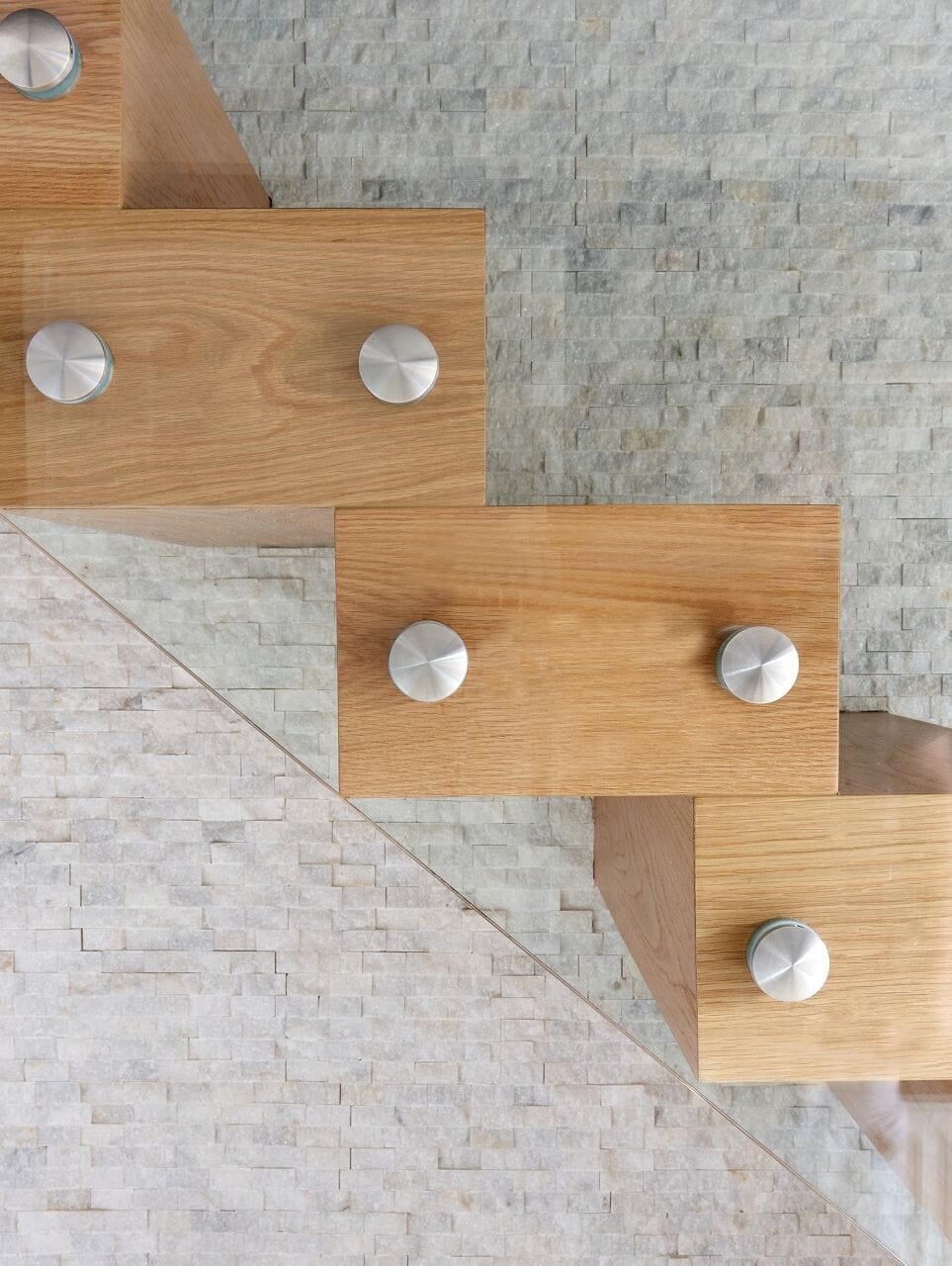 Detail of oak stair treads and round brushed-metal fixings holding a frameless glass balustrade against a stone feature wall.