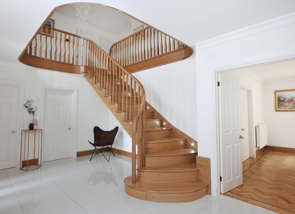 Curved wooden staircase with shaped timber spindles, illuminated steps and a matching gallery landing in a white hallway.