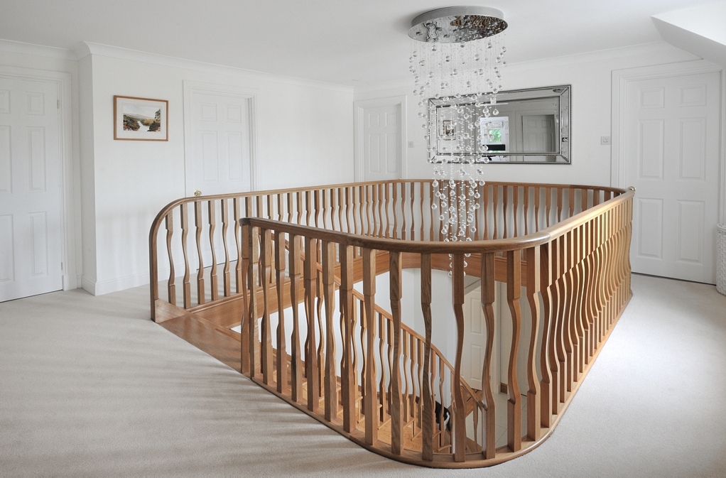 Curved oak gallery balustrade with shaped wooden spindles and a continuous handrail around an upstairs stairwell beneath a crystal chandelier.