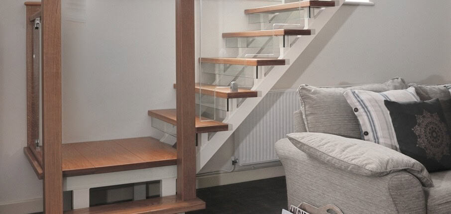 Open-riser staircase with wooden treads, white stringers, square timber posts and clamped glass panels beside a grey living-room sofa.