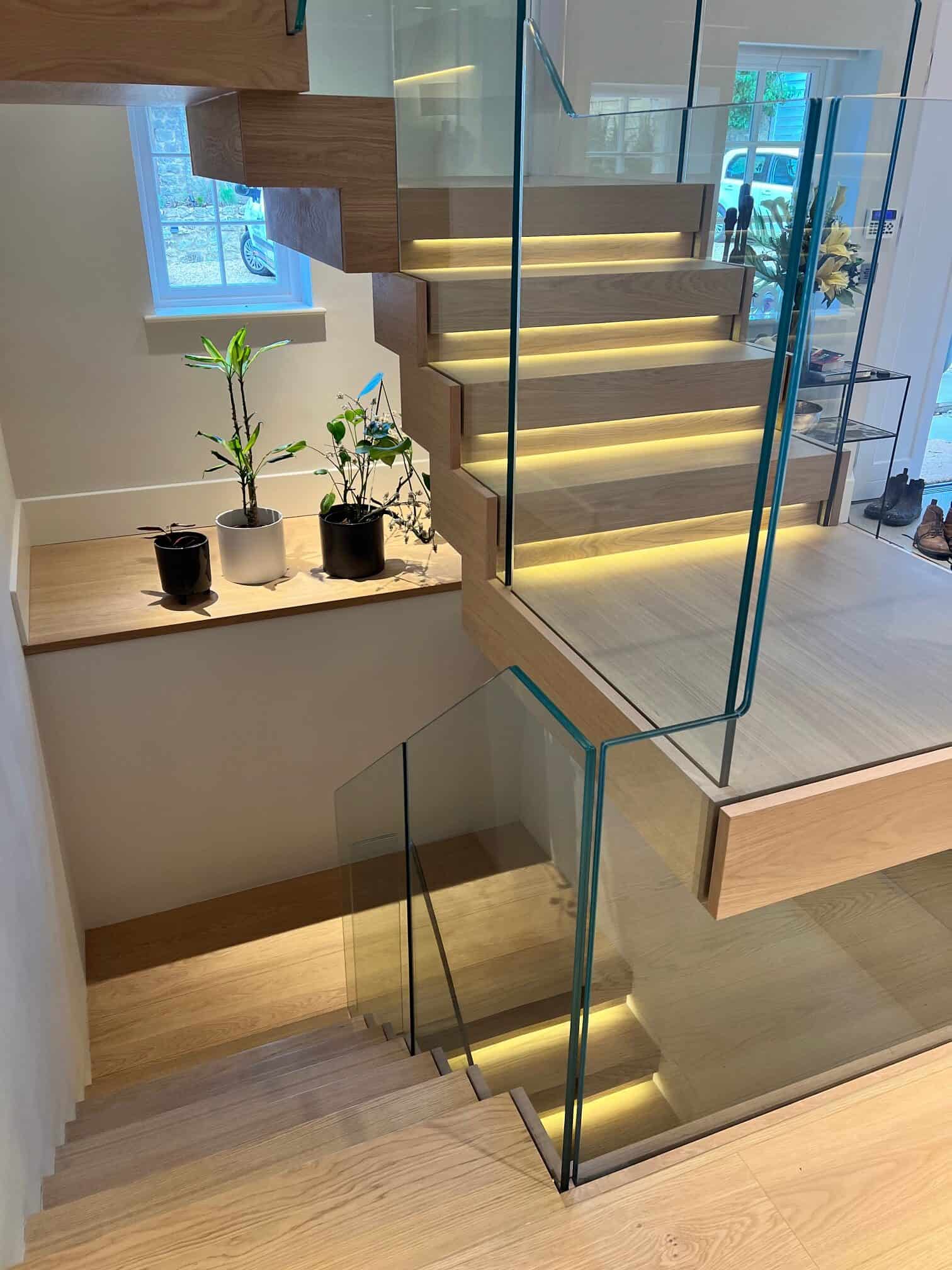 View across multiple flights of a pale wooden staircase with illuminated treads and shaped frameless glass balustrades.