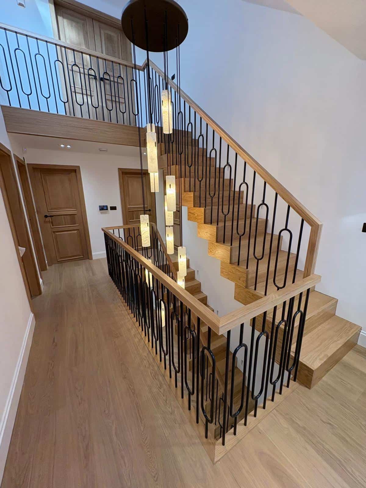 Oak Kubos staircase and gallery landing with black steel Arundel spindles forming elongated oval shapes beneath a cascade of pendant lights.