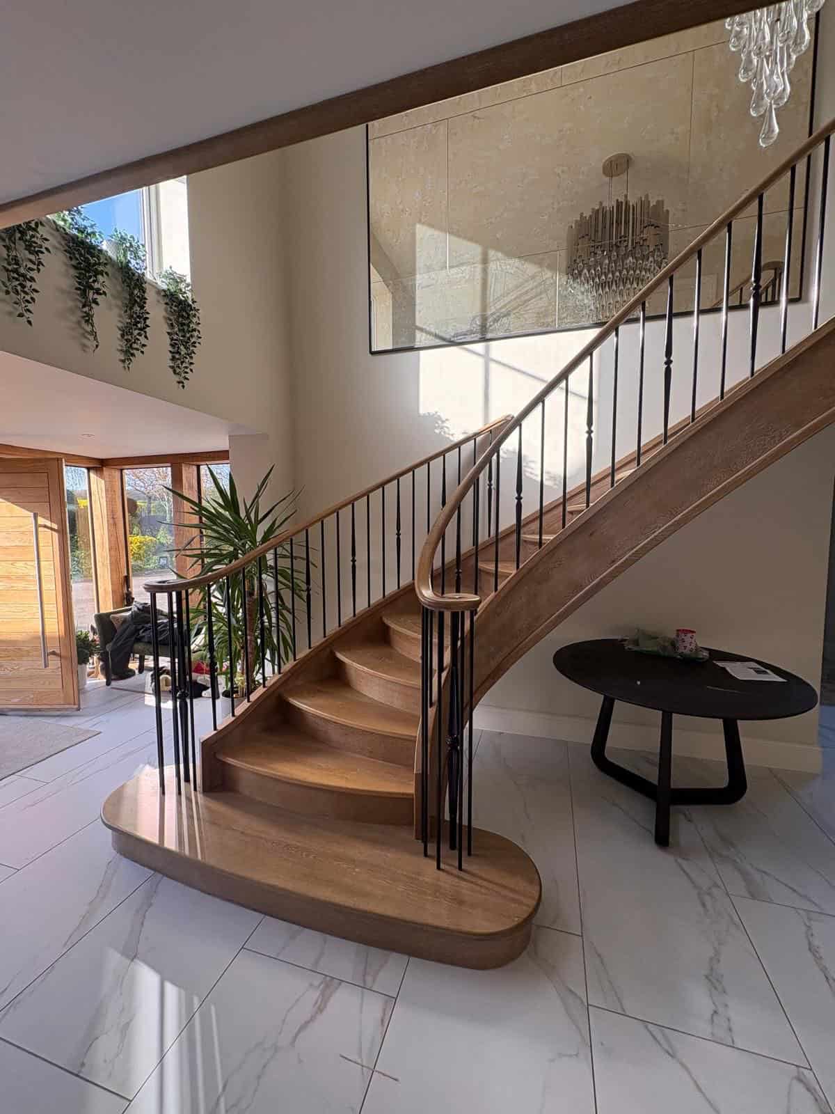 Curved oak helical staircase with bow-fronted treads, slender black metal spindles and a broad rounded bottom step in a bright marble-tiled hallway.