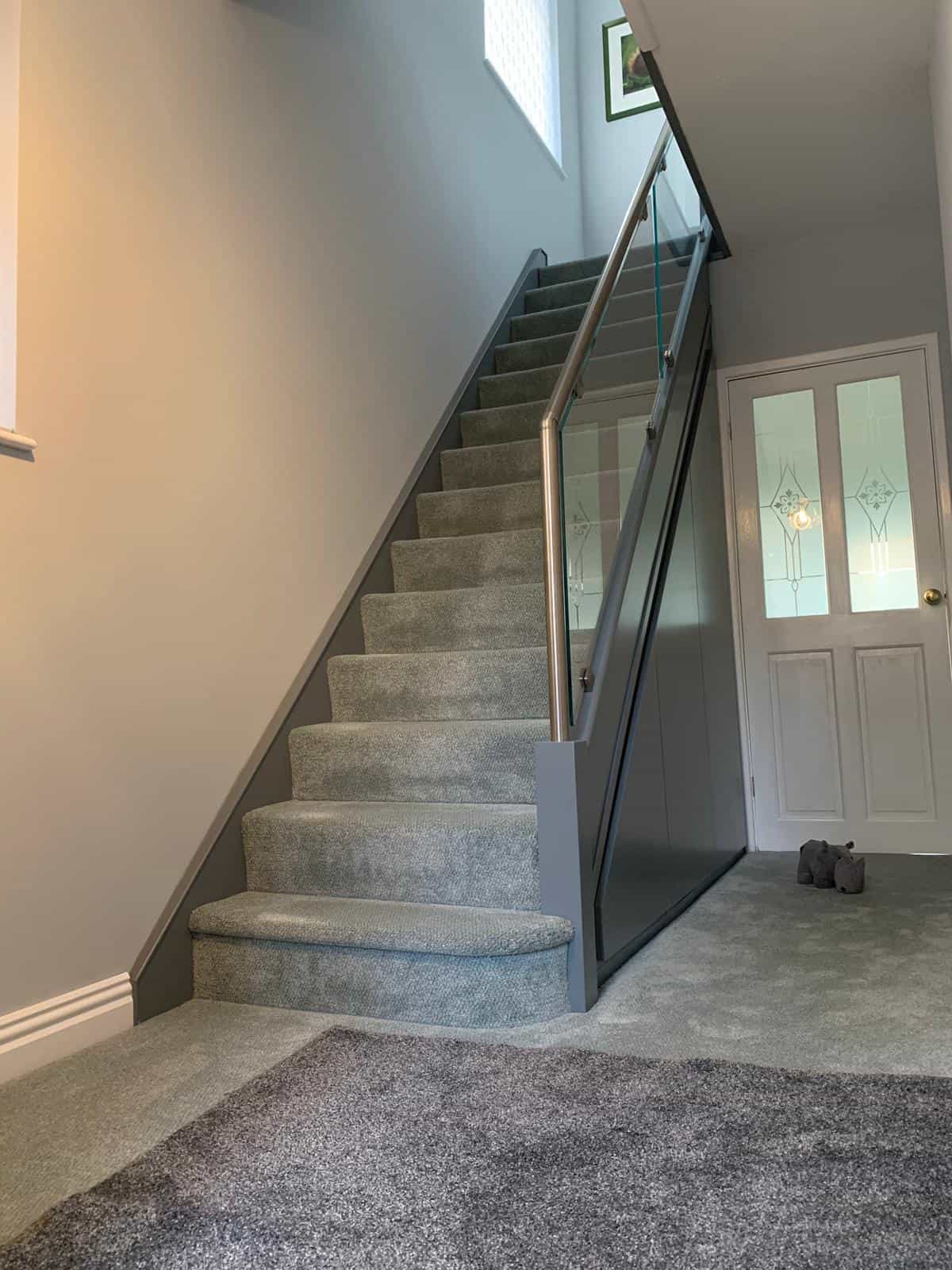 Grey carpeted staircase with a stainless steel handrail, clear glass balustrade and dark grey fitted cupboard doors beneath the stairs.