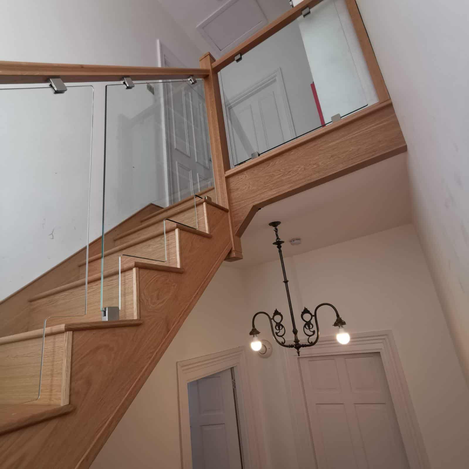 Oak staircase and upper landing with square wooden posts, matching handrails and clear glass balustrades secured by metal brackets.
