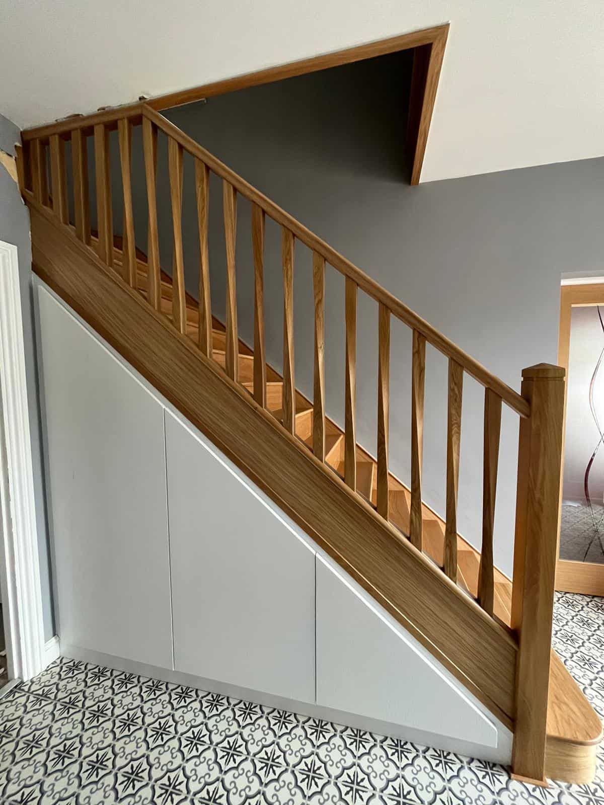 Oak staircase with slim twisted wooden spindles, square newel posts and white fitted cupboard doors beneath the stairs above patterned floor tiles.