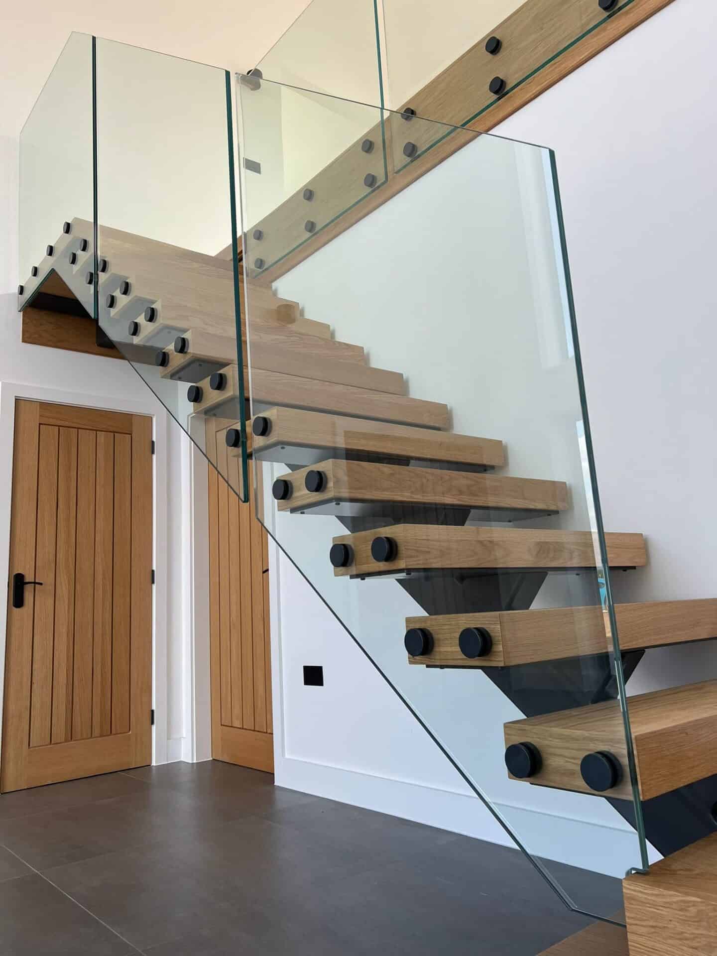 Open-riser oak staircase supported by a black central steel spine with frameless glass balustrades and round black fixings.