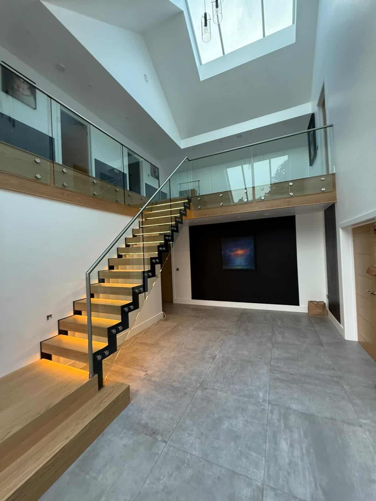 Illuminated oak open treads on black zigzag steel stringers with glass balustrades and a stainless steel handrail beneath a large skylight.