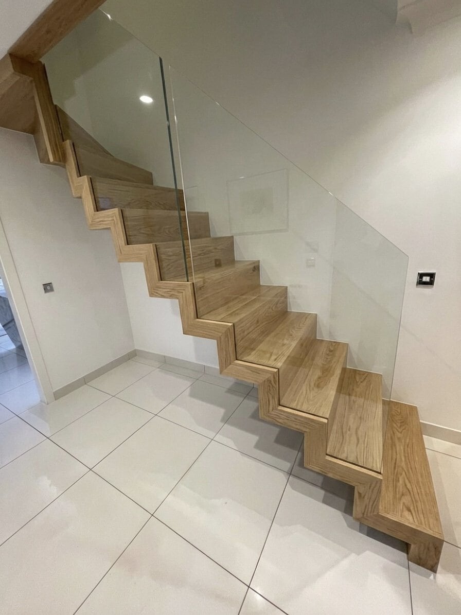Oak closed-tread zigzag staircase with a continuous stepped profile and frameless glass balustrades in a white tiled hallway.