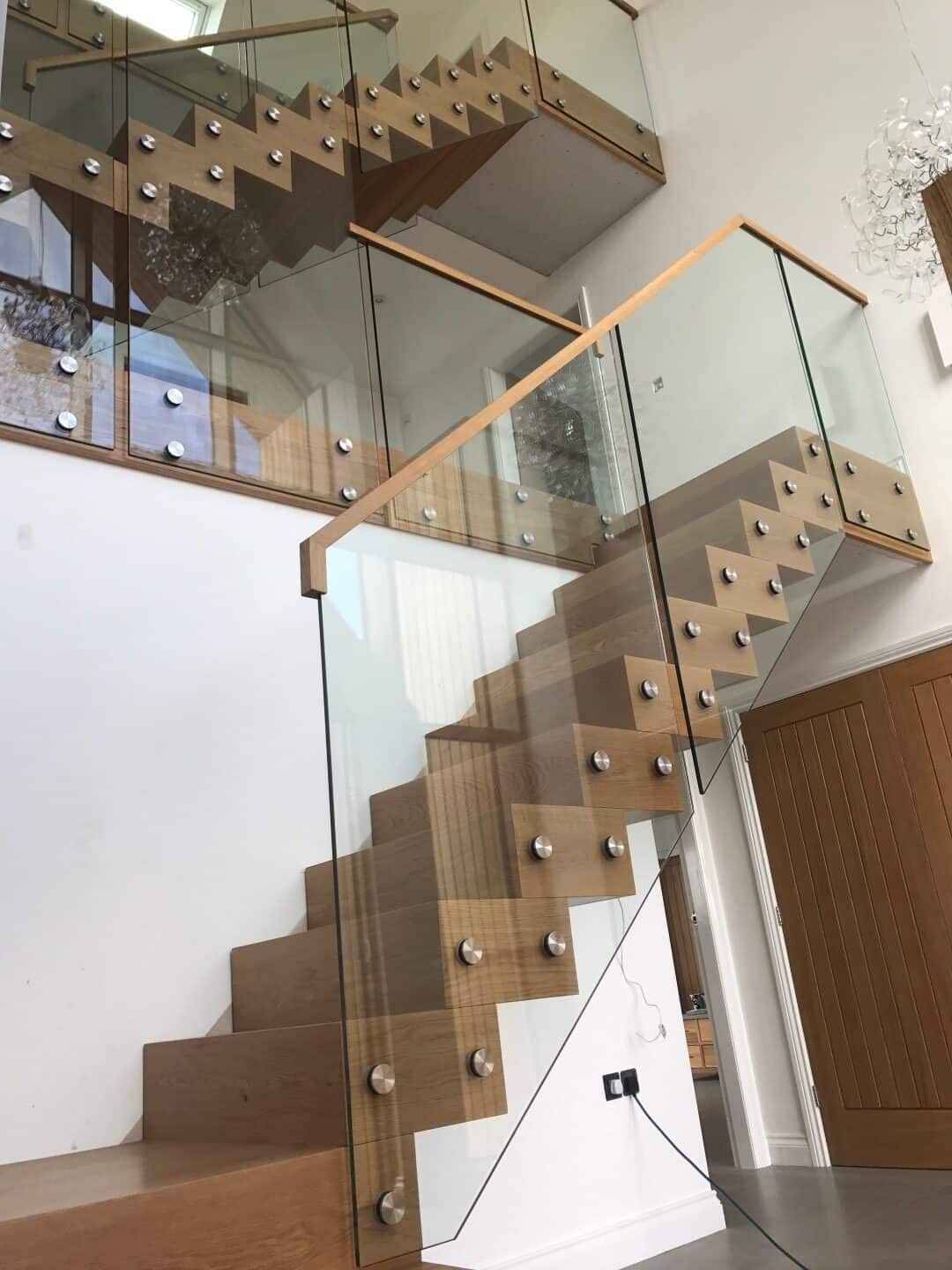 Kubos oak staircase with closed treads, zigzag edges and glass balustrades secured by round metal fixings, rising across several levels.