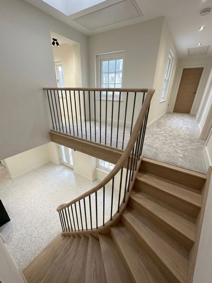 View down a natural oak helical staircase with slender black metal spindles and a continuous curved wooden handrail beside a carpeted upper landing.