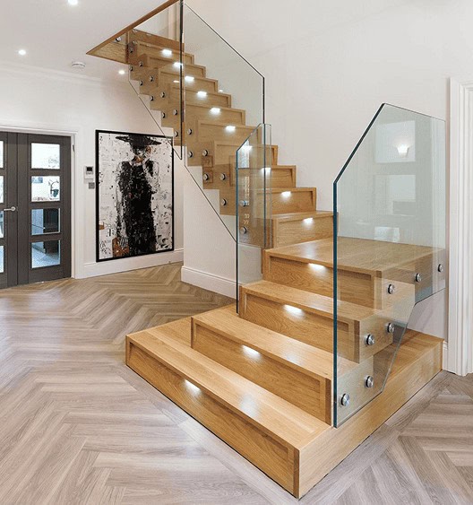 Oak staircase with recessed step lights, frameless glass panels secured by round metal fixings and broad rectangular feature steps above herringbone flooring.