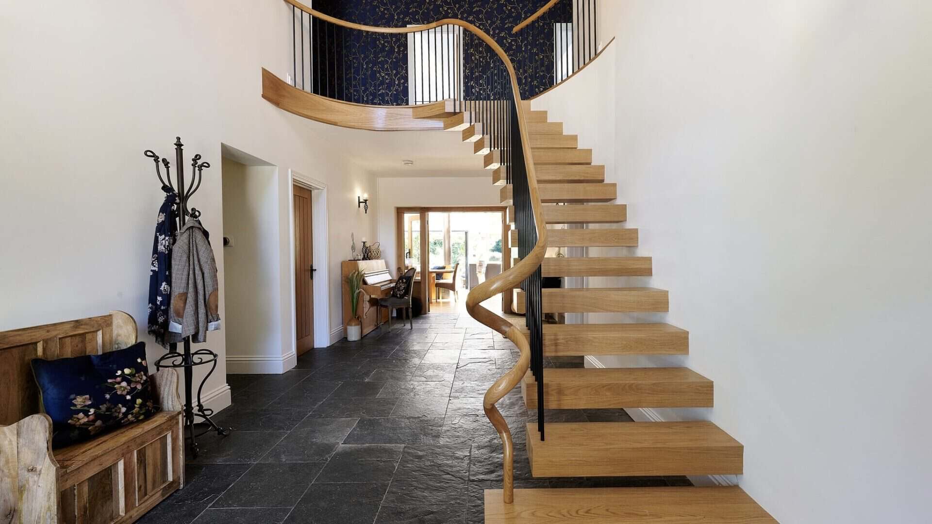 Helical floating oak staircase with open treads, black wrought iron spindles and a sweeping wooden handrail in a tiled entrance hall.