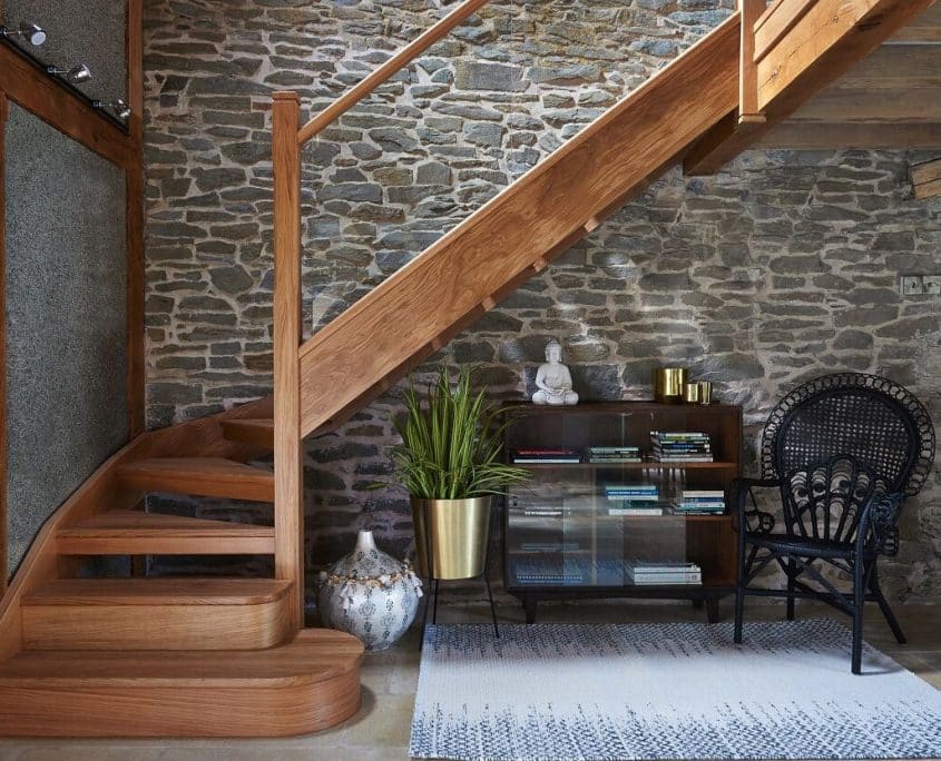 Oak open-riser staircase with glass balustrades and broad rounded wooden feature steps beside an exposed stone wall in the Morris family home.