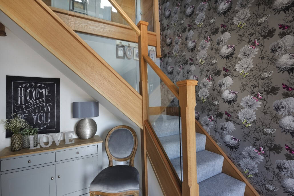 Oak staircase with glass balustrades, square newel posts and grey carpet beside a floral feature wall.