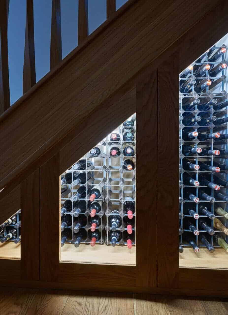 Illuminated wine racks behind glass-fronted wooden doors fitted beneath an oak staircase.