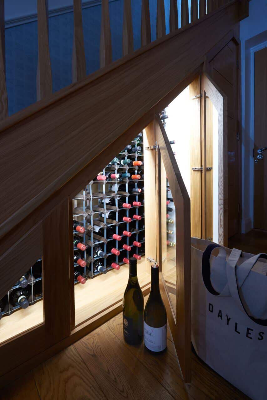 Open glass-fronted door revealing illuminated wine racks built into the space beneath a wooden staircase.