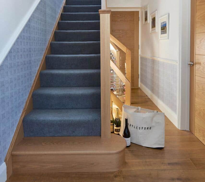 Blue carpeted staircase with oak newel posts, a rounded oak bottom step and illuminated wine storage beneath the stairs.