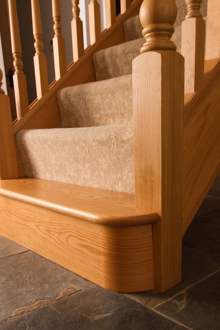 Close-up of a rounded oak bottom step beside a square newel post on a traditional staircase with beige carpet and turned wooden spindles.