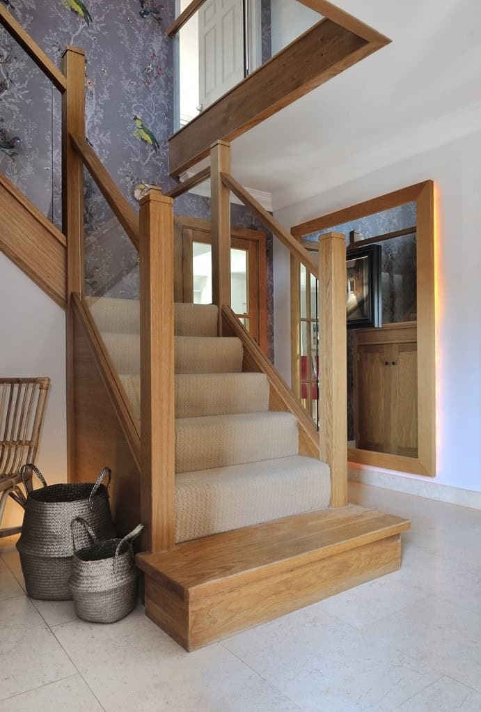 Oak staircase with glass balustrades, cream carpeted steps and a broad rectangular oak feature step in a tiled hallway.
