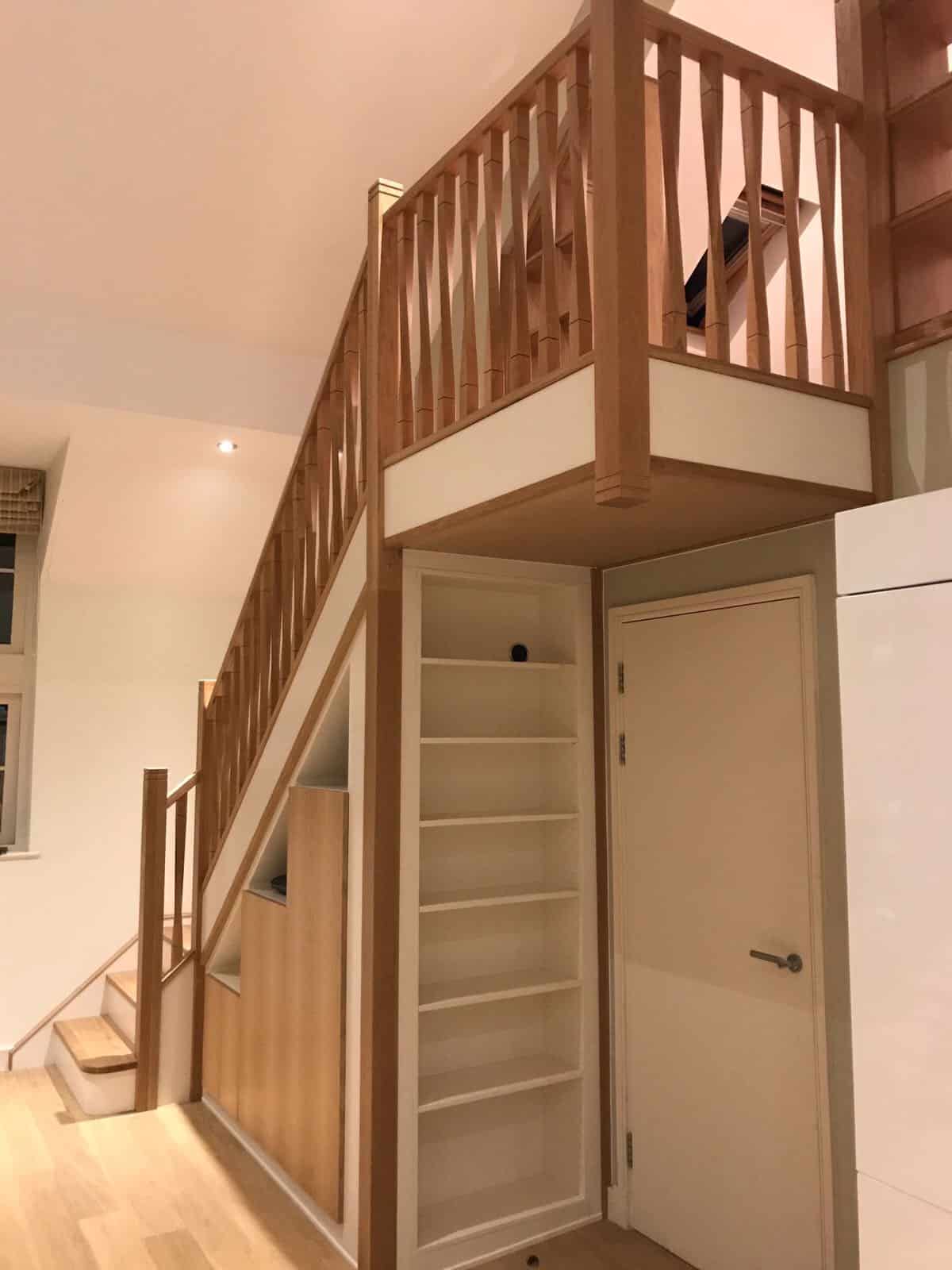 Oak staircase and landing with square wooden spindles, fitted cupboards beneath the stairs and tall open shelving beside a doorway.