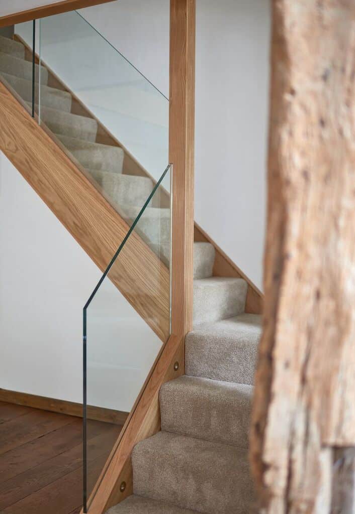 Detail of a carpeted turning oak staircase with glass balustrades and a continuous wooden handrail beside an exposed timber post.