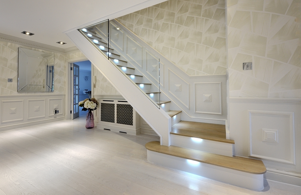 White painted staircase with oak treads, recessed step lights and frameless glass balustrades above broad rounded feature steps in a panelled hallway.