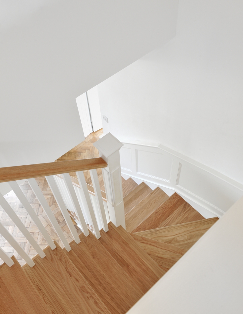 View down a turning staircase with natural oak treads and handrail, white square spindles and a white box newel post beside a panelled wall.