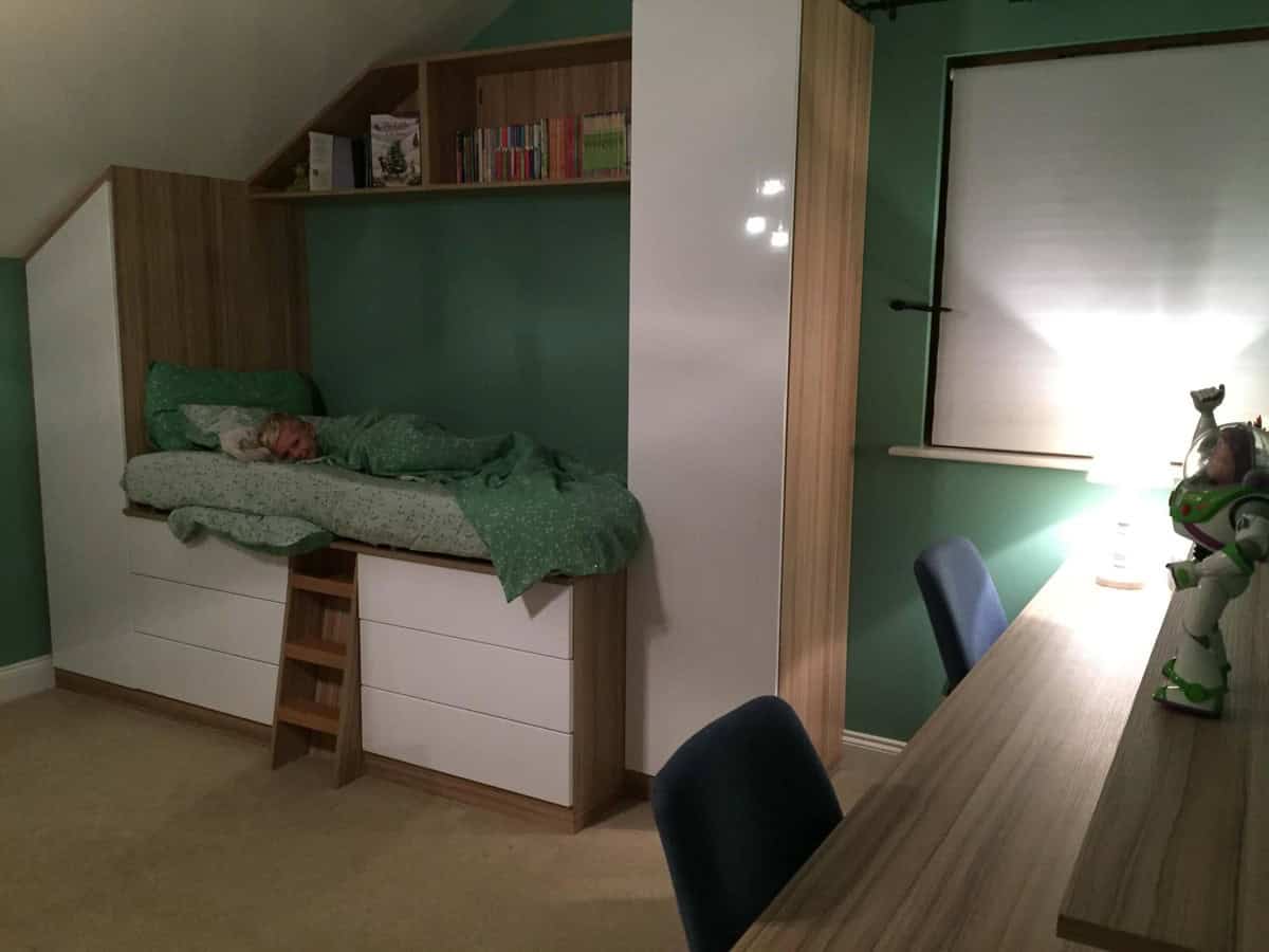 Fitted bedroom with a raised bed above white storage drawers, a small wooden ladder, overhead bookshelves and a long desk beneath a sloping ceiling.