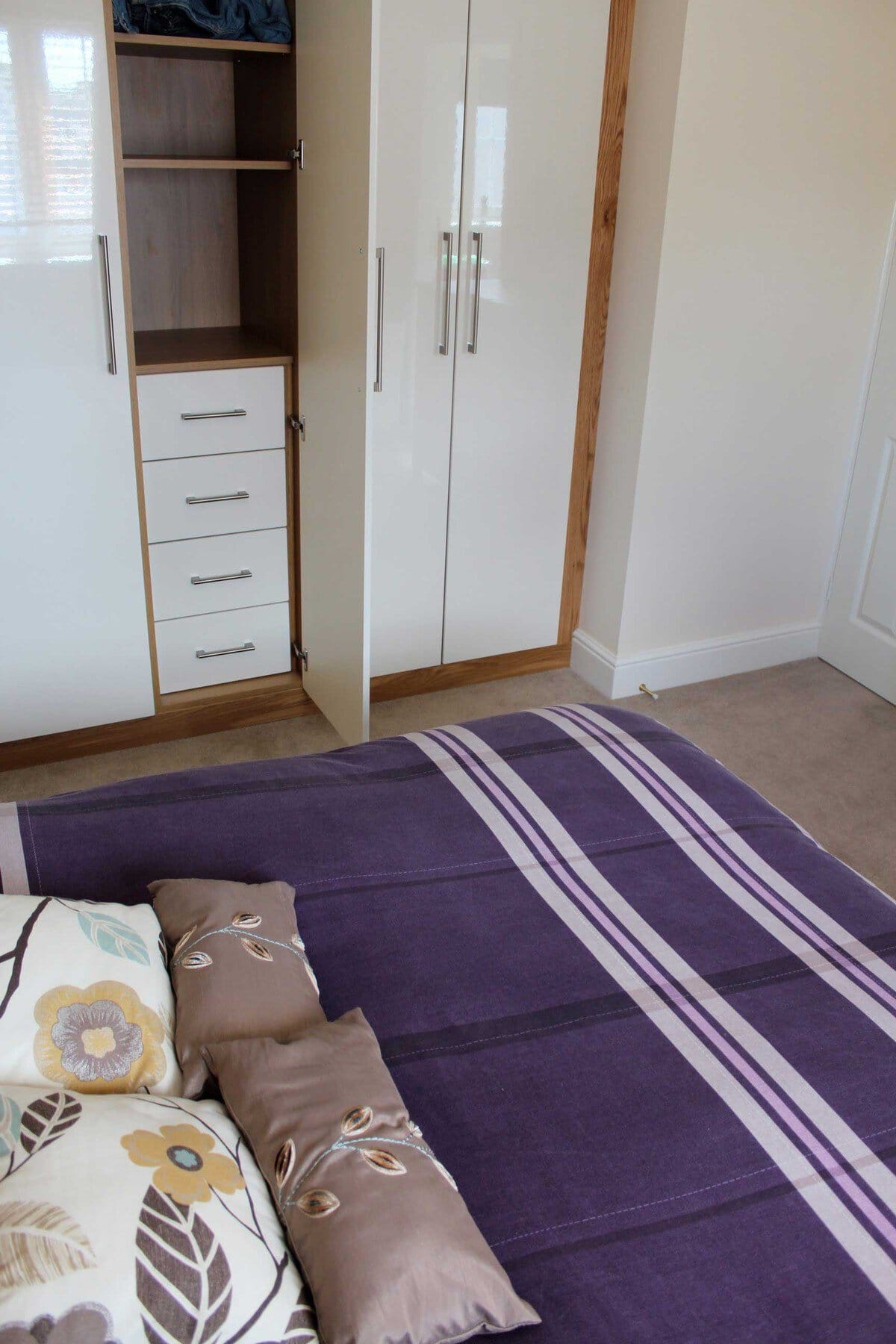White gloss fitted bedroom wardrobes with oak trim, long metal handles and an open door revealing shelves and a stack of drawers beside a purple bedspread.