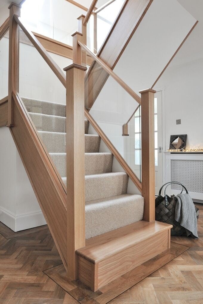 Carpeted oak staircase with integrated glass panels, tall square newel posts and a rectangular wooden feature step above parquet flooring.